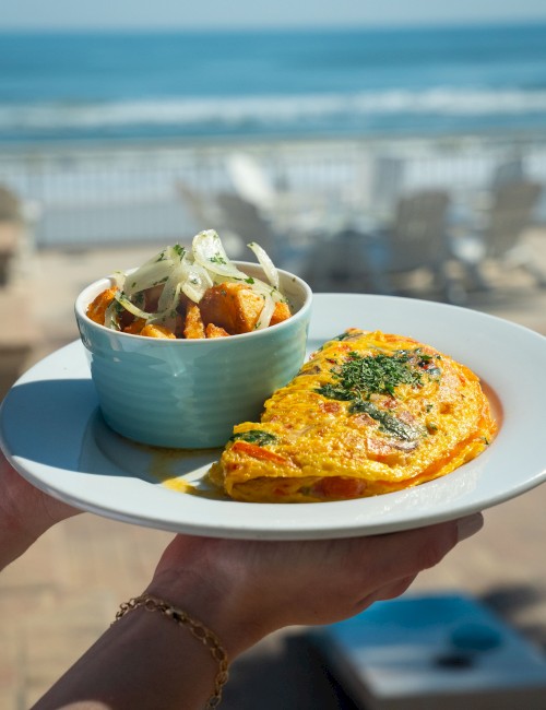 A plate with an omelette topped with herbs, alongside a bowl of salad or mixed vegetables, with a seaside background.
