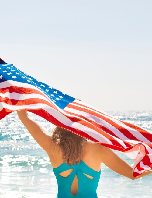 A person in a swimsuit stands by the sea, holding an American flag above their head in a bright, sunny setting.