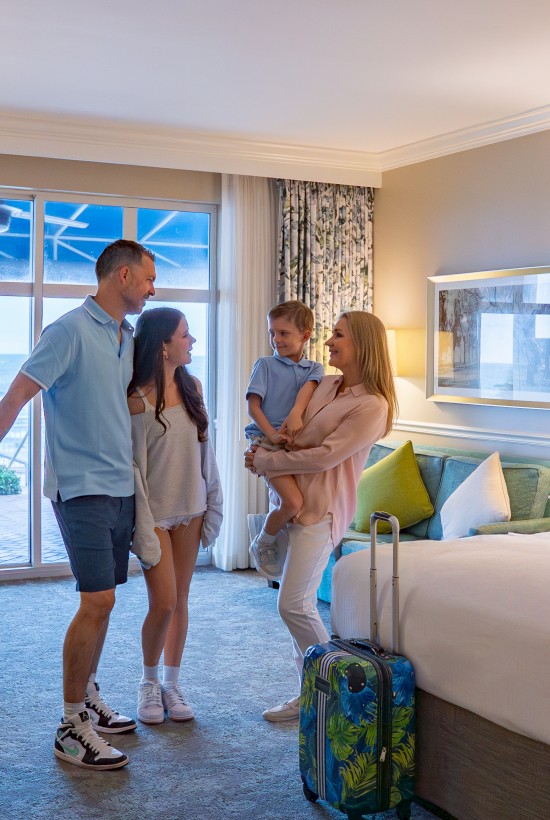 A family of four stands in a hotel room with luggage, enjoying the sea view through the window.