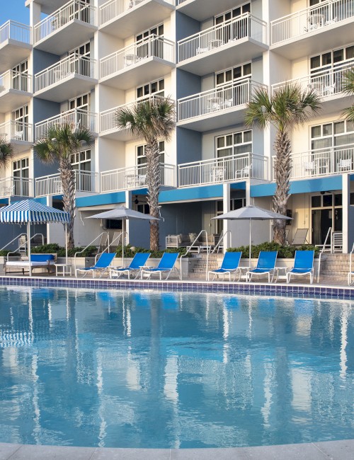 A hotel with blue balconies and a pool surrounded by blue lounge chairs and umbrellas, set under clear skies.
