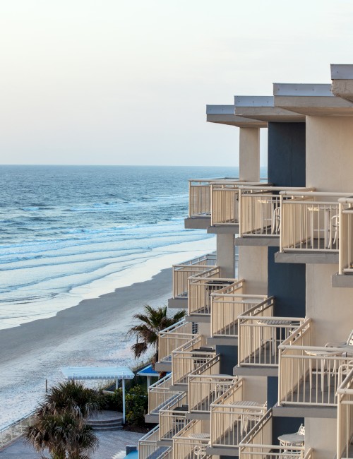 The image shows a beachfront hotel with multiple balconies overlooking the ocean, with palm trees visible below on the sandy beach area.