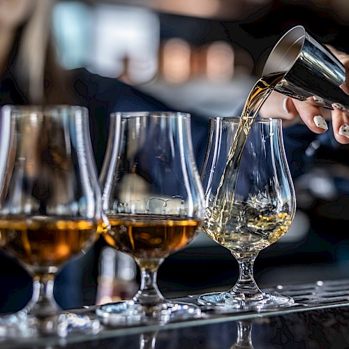 A person is pouring amber liquid from a jigger into a glass, alongside several similar glasses on a bar counter.