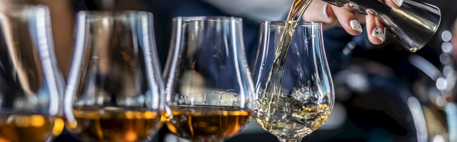 A person is pouring a drink into a row of glasses, likely spirits, in a bar setting.