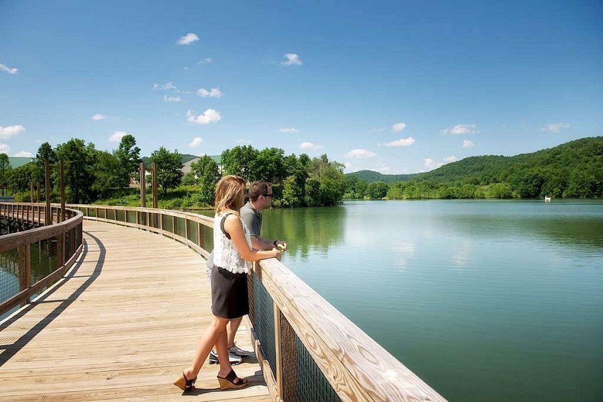 A couple stands on a wooden boardwalk over a serene lake, surrounded by lush greenery and hills under a clear blue sky.