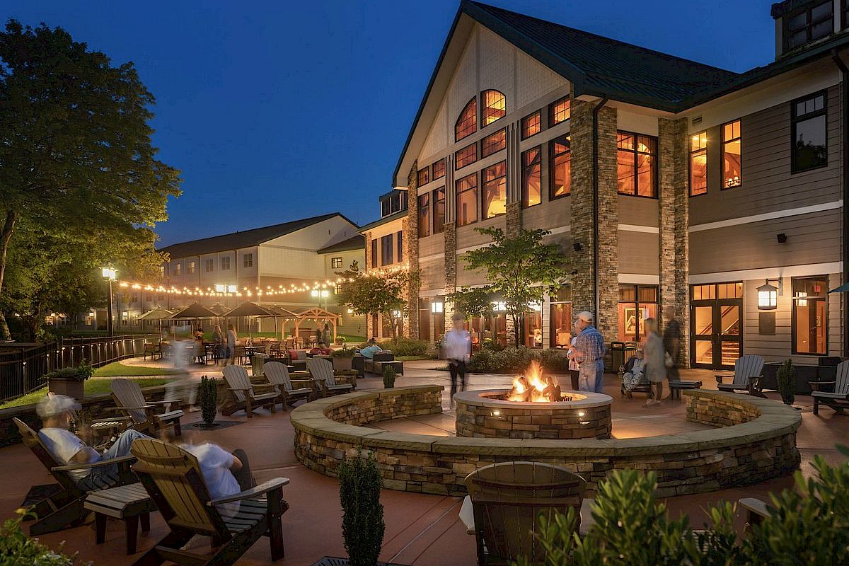 The image shows a cozy outdoor patio area of a building at twilight. People are gathered around a fire pit with string lights overhead, creating a warm ambiance.