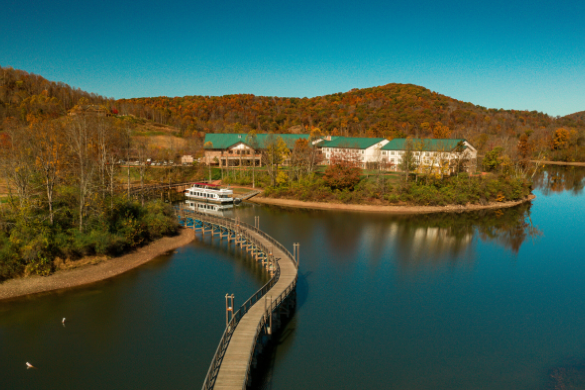 An aerial view shows a winding bridge leading to buildings surrounded by trees, with a boat docked nearby on a calm lake, under a clear sky.