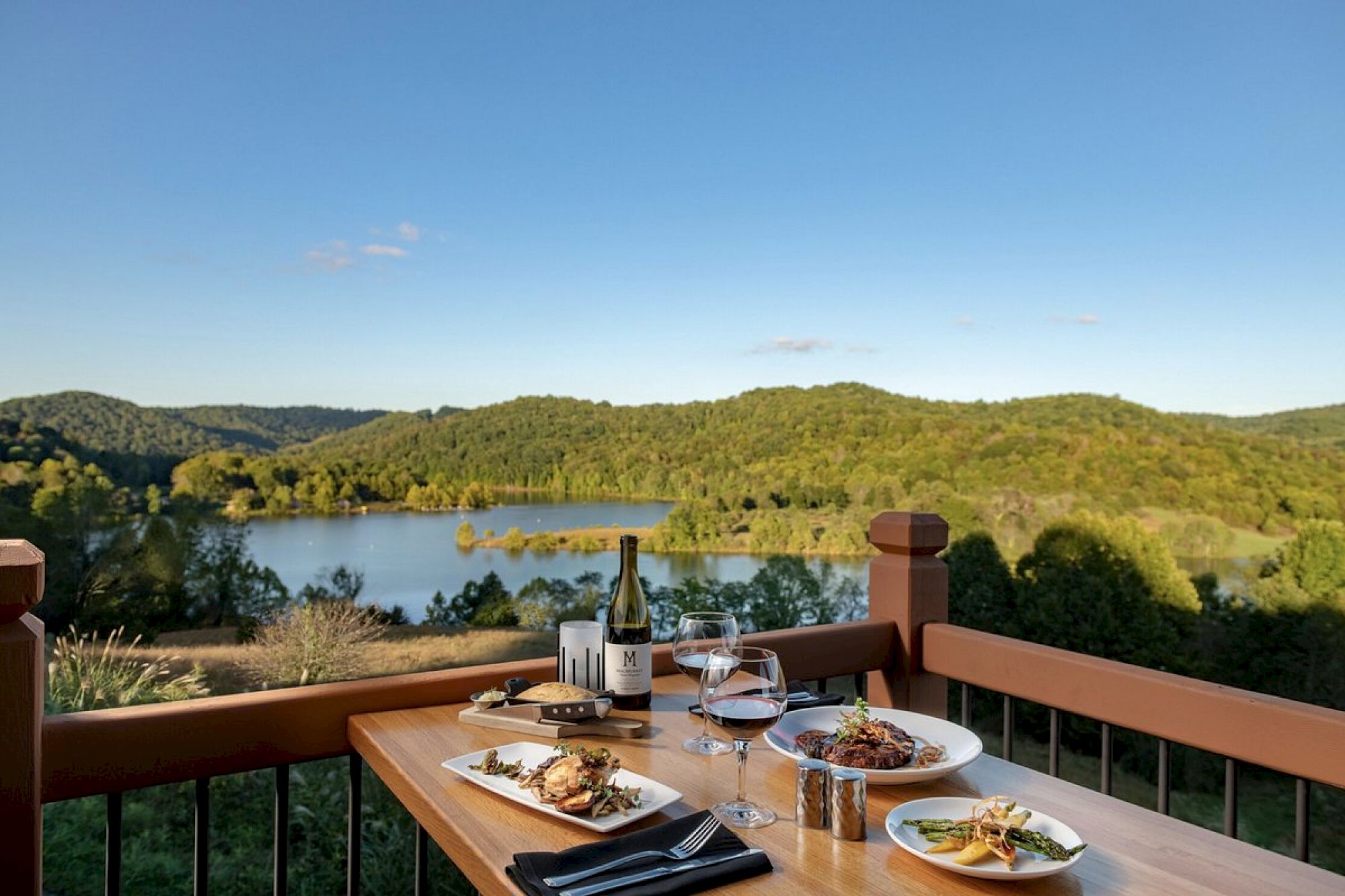 A table set with food and drinks overlooks a scenic lake and lush green hills under a clear blue sky, creating a picturesque dining experience.