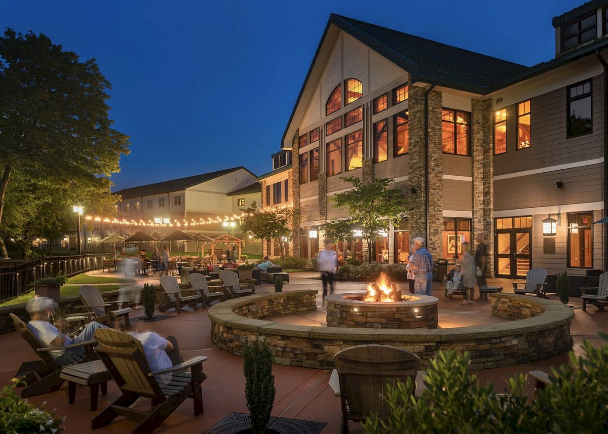 An outdoor lounge area with a fire pit, surrounded by chairs, festive lights, and a large building in the background during the evening.