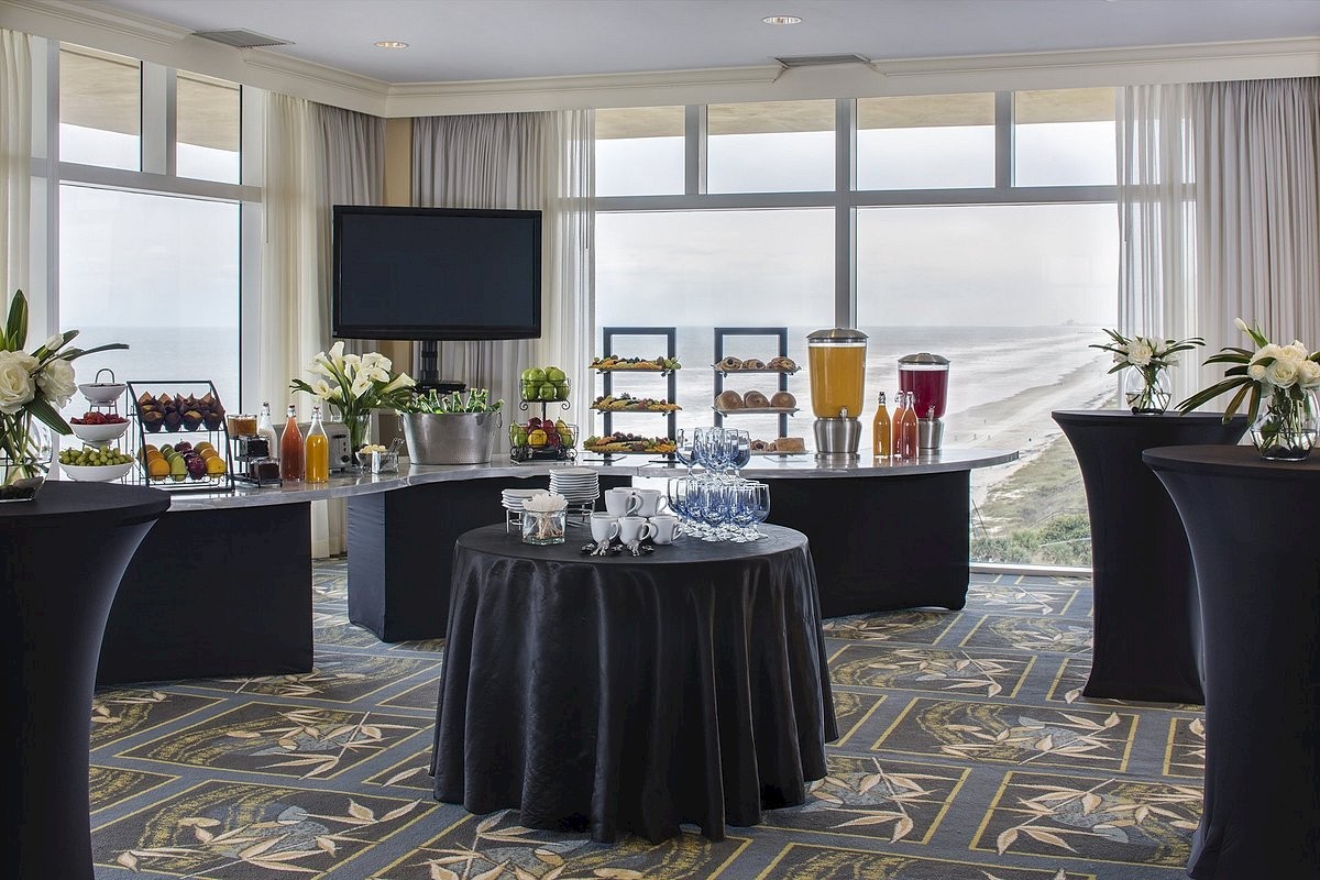 A bright hotel breakfast setup with high-top tables, a central round table, juice dispensers, pastries, flowers, and a wall-mounted TV, by large windows.