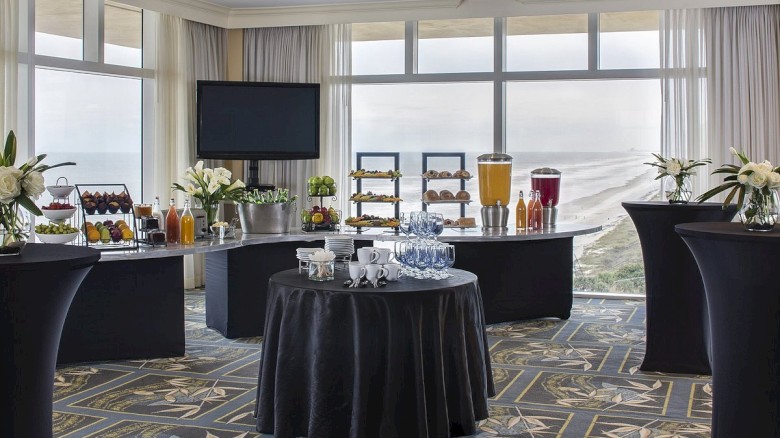 A bright hotel breakfast setup with high-top tables, a central round table, juice dispensers, pastries, flowers, and a wall-mounted TV, by large windows.