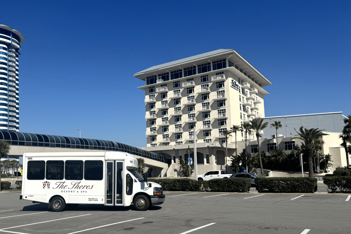 A white shuttle bus with “The Gables” or similar text on the side sits in a parking lot, in front of a large multi-story hotel with balconies, palm trees, and a clear blue sky.