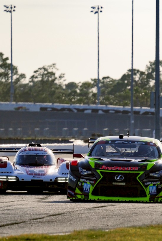 Two race cars sprint side-by-side on a track, slicks blazing, with a blurred grandstand and tall light poles in the background. End.