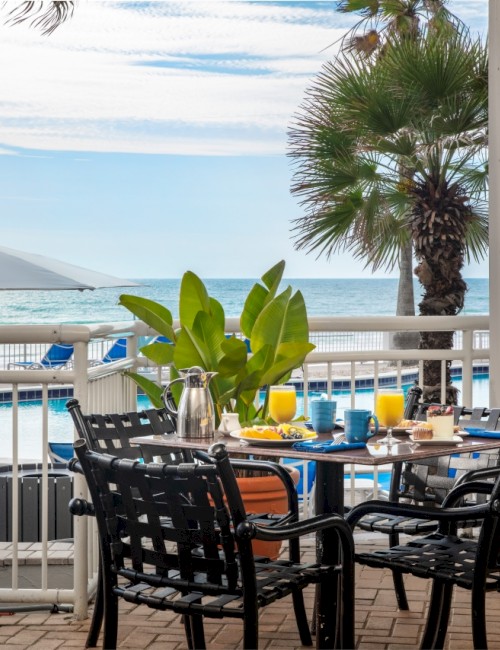 A seaside restaurant terrace with black wicker chairs, a few tables set with yellow and blue items, tropical plants, palm trees, and a view of the ocean under a bright sky.