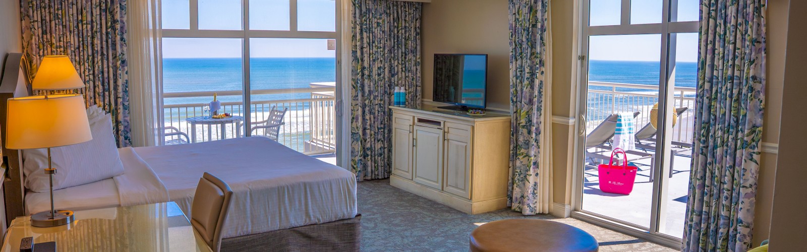 A hotel room with ocean view features a bed, desk, TV, seating area, and a balcony with chairs, overlooking the sea under a clear sky.