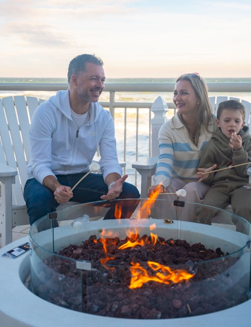A family sits around a glowing fire pit on a deck, chatting and smiling at sunset, with a baby in blue and white striped sweater nearby.