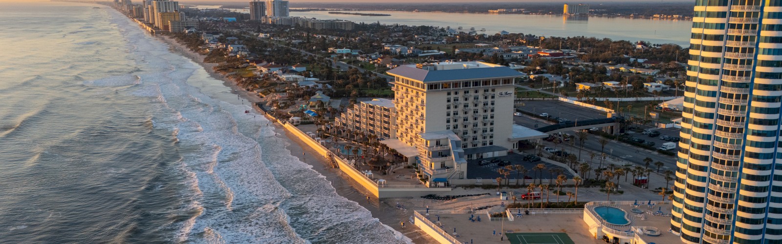 Aerial view of a coastal city at sunset with a long beach, waves, high-rise buildings, a waterfront promenade, and a small green sports area near the shoreline.