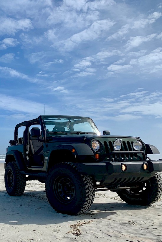 A black off-road Jeep with large tires on a sandy beach, waves in the background, sunny blue sky, open-top adventure vibes.