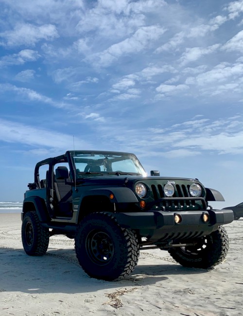 A black open-top Jeep Wrangler sits on a wide, sandy beach with the ocean in the background and a bright, partly cloudy blue sky.