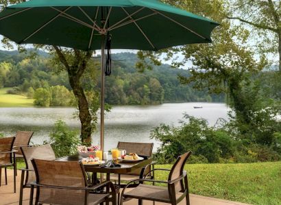 An outdoor patio with a green umbrella-covered table set for a meal, overlooking a serene lake surrounded by lush greenery and hills in the background.