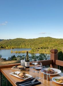 A scenic outdoor dining scene overlooking a lake with green hills. The table has plates of food, wine, glasses, and utensils, ending the sentence.