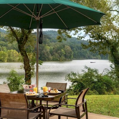 An outdoor dining setup with a table, chairs, and a green umbrella overlooking a serene lake with hills and trees in the background.