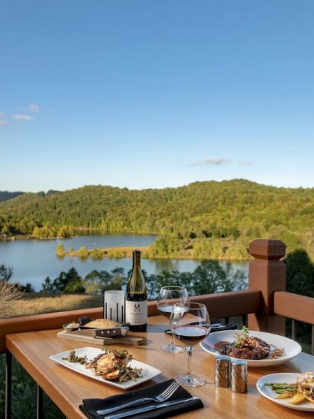 A dining table set with meals and wine overlooks a scenic lake and lush green hills under a clear blue sky, providing a picturesque view.