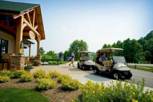 The image shows two people in golf carts near a building with a green roof, while another person in the background walks holding a golf bag.