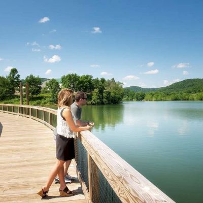 Two people are standing on a wooden boardwalk overlooking a scenic lake with surrounding greenery and hills under a clear blue sky.