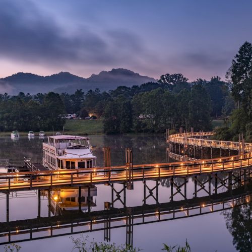 A serene landscape features a lit curved wooden pier over still water, a boat docked, lush trees, and mountains under a twilight sky.