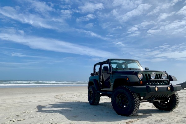 A black off-road Jeep on a sandy beach with the ocean and blue sky in the background, rugged tires and open-top driving vibe.