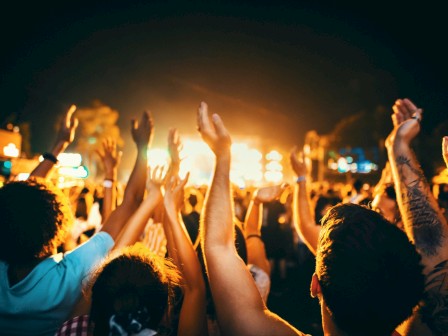 A lively concert crowd raises their hands toward a bright stage under warm lights, silhouettes dancing as the music cues the night.