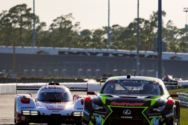 Two race cars chase on a track, a Lexus leads with bright green accents while another white-and-red car stays close behind.