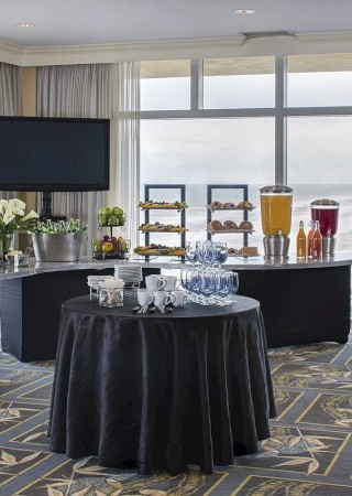 A hotel conference room set up for a buffet: several high and round black tables with white flowers, beverage dispensers, juice, pastries, and a TV on the wall, all by large windows.