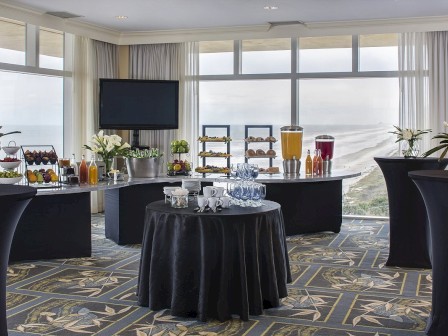 A hotel conference room set up for a buffet: several high and round black tables with white flowers, beverage dispensers, juice, pastries, and a TV on the wall, all by large windows.