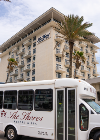 A resort building with a shuttle bus parked in front, surrounded by palm trees, under a partly cloudy sky.