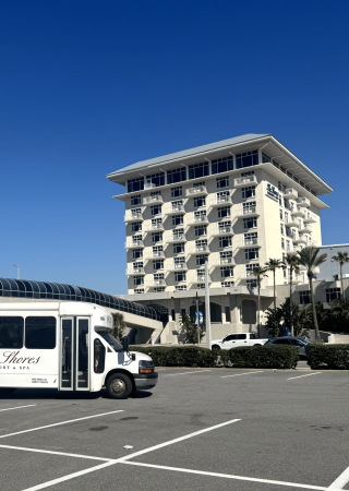 A white shuttle bus in a parking lot with a modern hotel building and blue sky behind it.