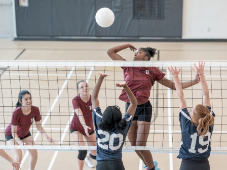 A group of girls playing volleyball indoors, one player jumps to spike the ball while teammates159 line up near the net, eager for the block.