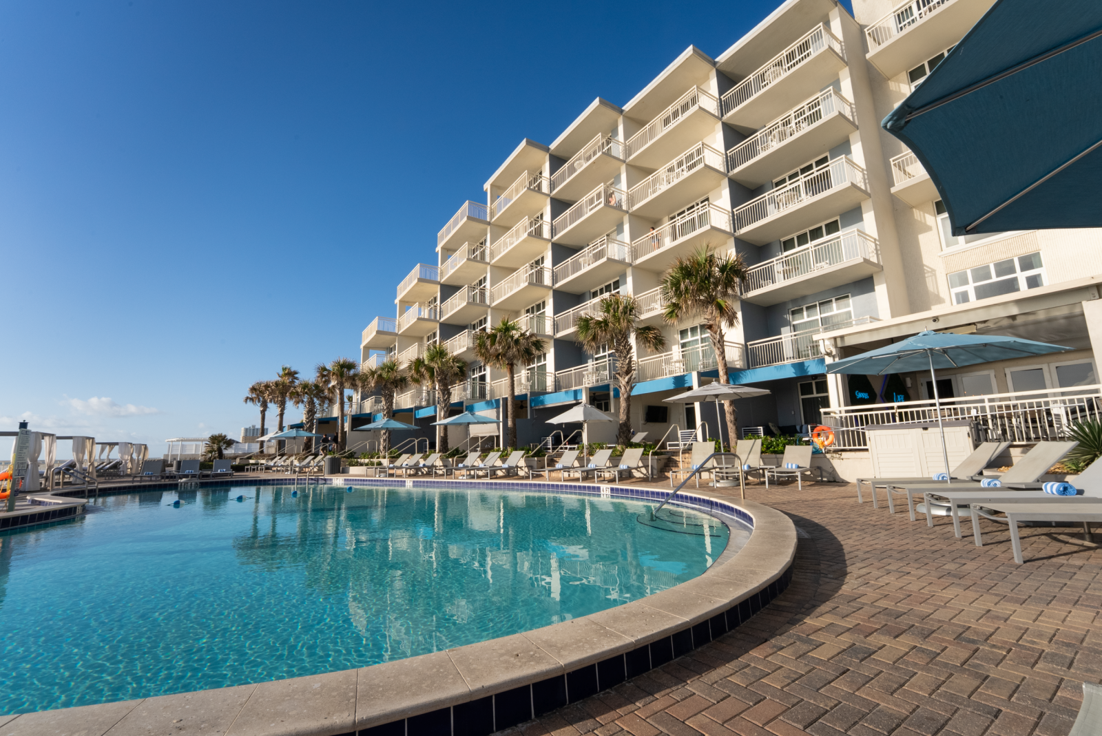 A sunny pool area with lounge chairs, umbrellas, and palm trees outside a modern hotel building under a clear blue sky.