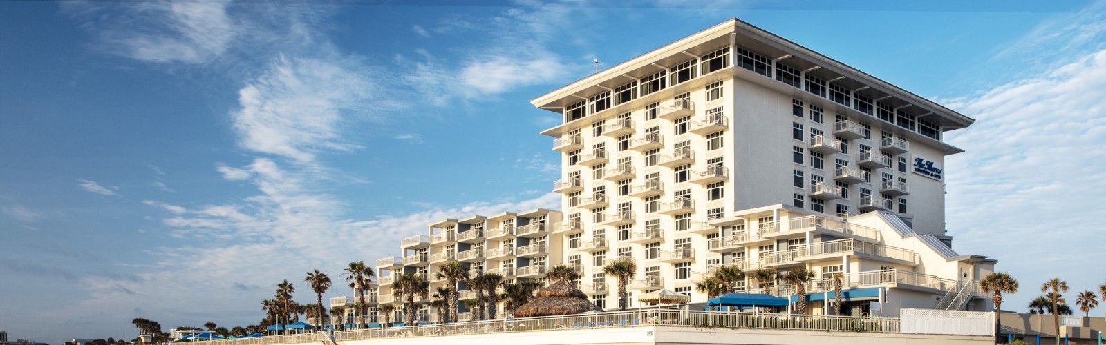 The image depicts a large multi-story hotel or resort building near a beach, with its reflection visible in the water. Ending the sentence.