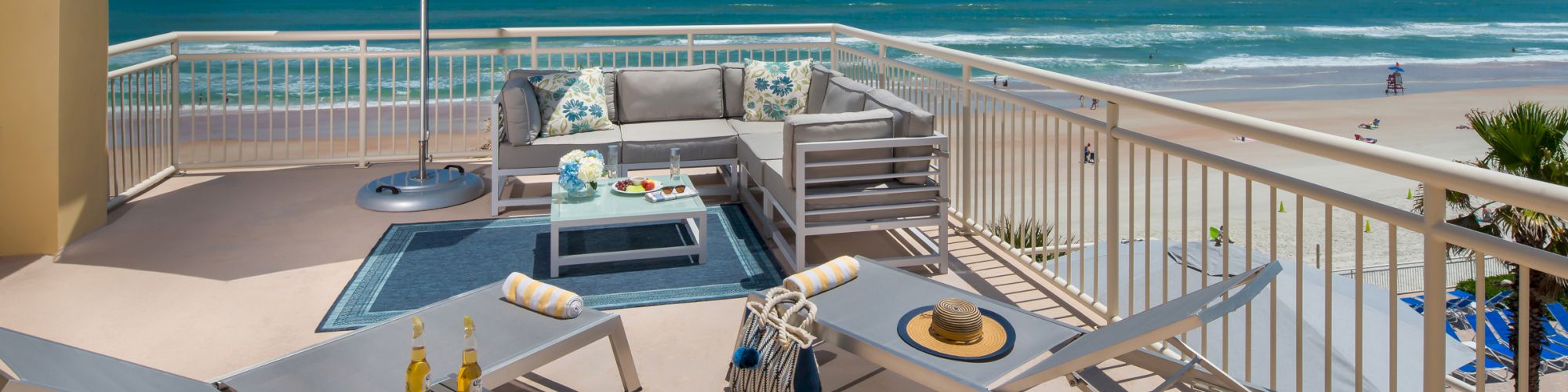 A beachfront patio with lounge chairs, an umbrella, a table, and drinks on a raised platform. Overlooks a beach and ocean on a sunny day.