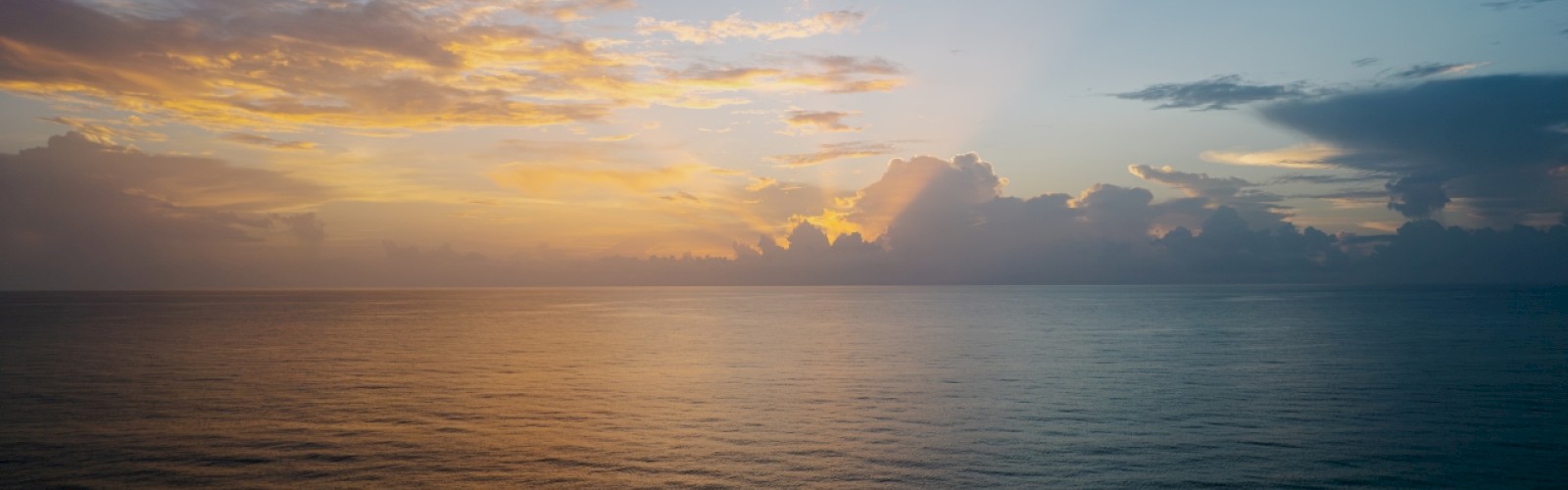 A serene ocean view at sunrise or sunset, with a colorful sky and clouds casting reflections on the calm water.