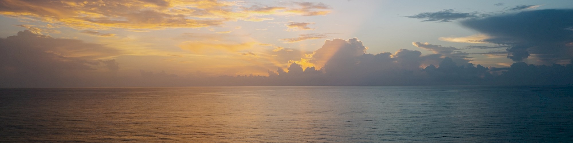 A serene ocean view at sunrise or sunset, with a colorful sky and clouds casting reflections on the calm water.