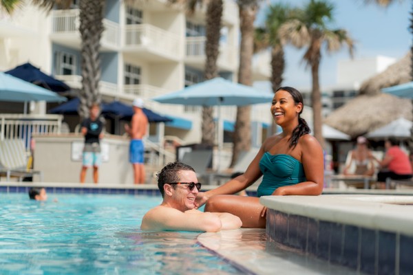 Two people enjoy a poolside moment, one in the water and one sitting on the edge, with a background of umbrellas and palm trees.