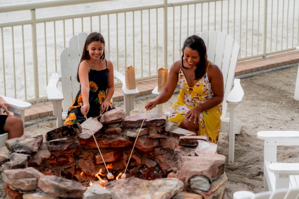 Two people roasting marshmallows over a stone fire pit, seated in white Adirondack chairs with sandy surroundings and a fence in the background.