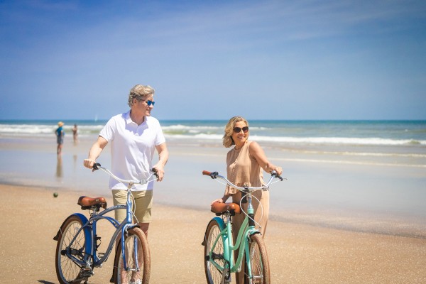 A man and a woman are walking with bicycles on a beach. The sky is clear and the sea is calm, making for a pleasant day by the shore.