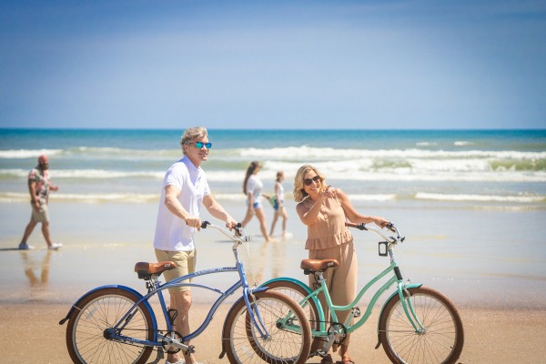 Two people stand with bicycles on a sunny beach, with the ocean in the background and a few others walking by.