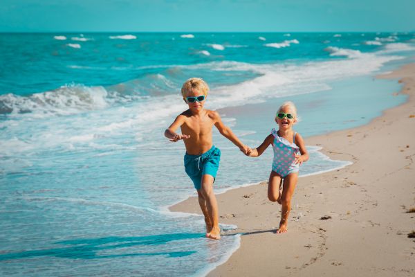 Two children wearing swimsuits and sunglasses run along a beach while holding hands, with waves gently crashing in the background.