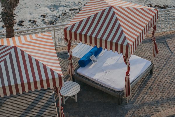 The image shows striped red and white beach canopies by a sandy beach, with lounge chairs and a nearby circular pool under a blue sky.