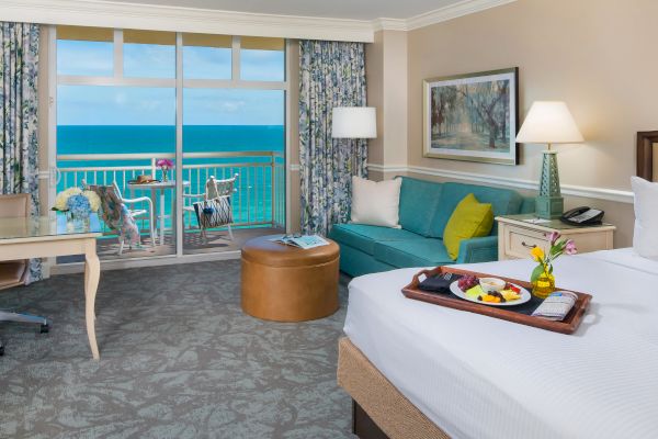 A hotel room with a bed, sofa, desk, and a balcony overlooking the ocean. There’s a tray with food on the bed and a chair on the balcony.