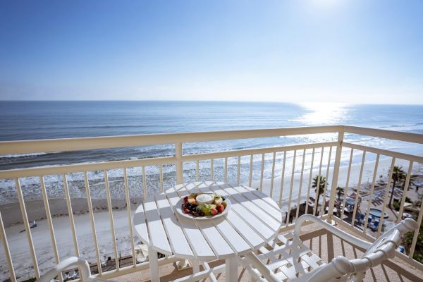 A balcony overlooking a serene, sunlit beach with a white table and chairs, featuring a bowl of fruit on the table.